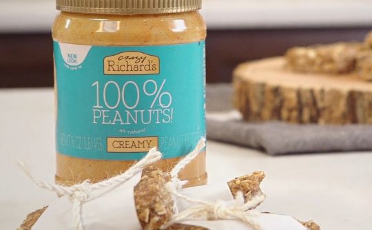 homemade granola bars wrapped in parchment paper tied with string, in front of a jar of Crazy Richard's creamy peanut butter