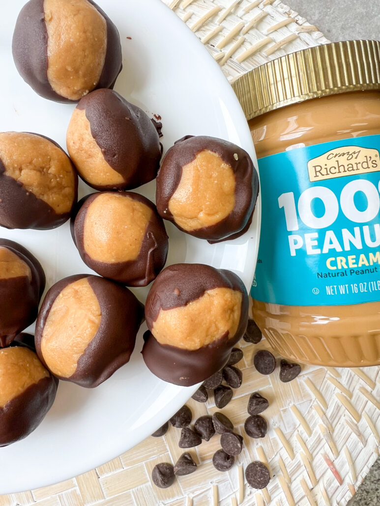 buckeyes (peanut butter balls dipped in chocolate) on a white plate, next to a jar of Crazy Richard's creamy peanut butter, a handful of chocolate chips sprinkled on a woven rattan placemat below.