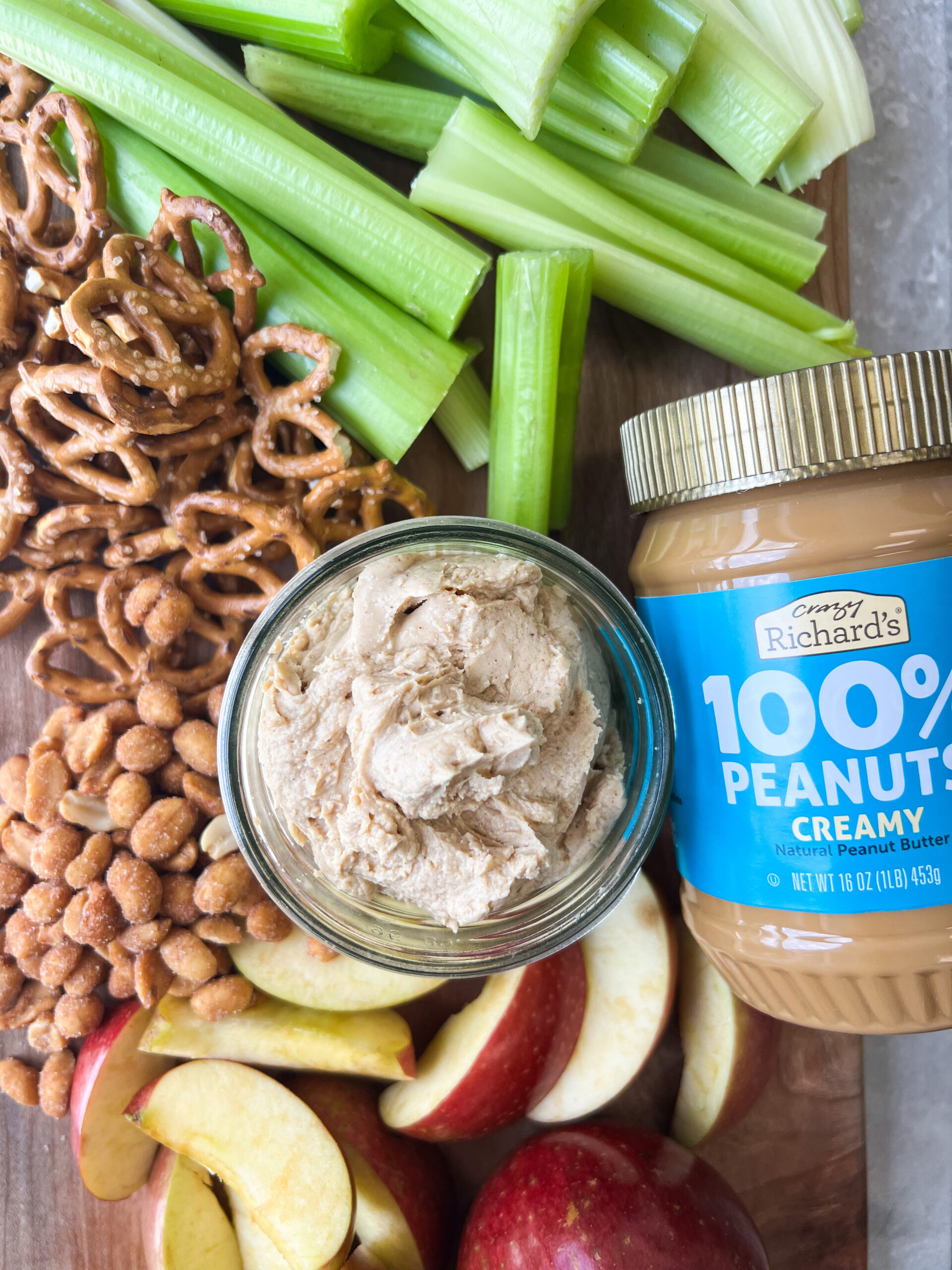 a snack board with celery, peanuts, pretzels, apple wedges, and peanut butter dip, and a jar of Crazy Richard's creamy peanut butter