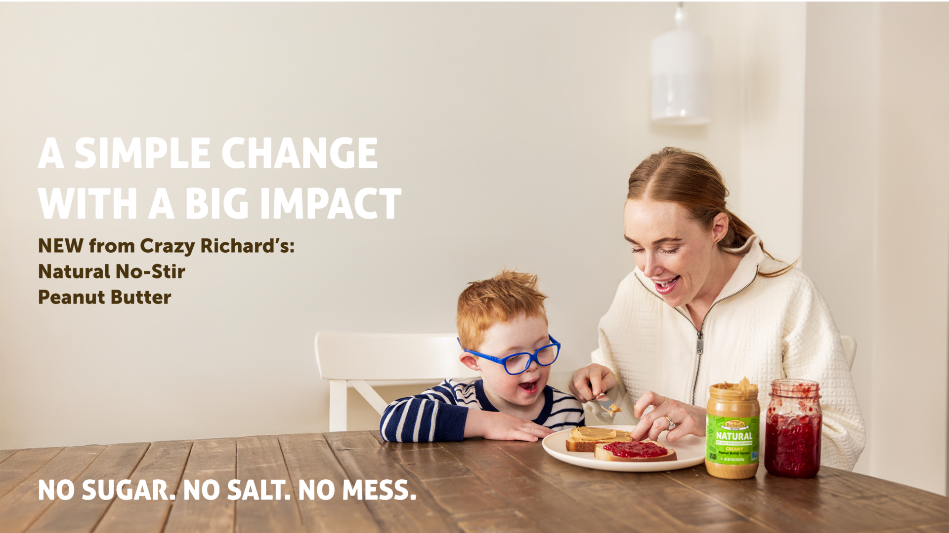 photo of a mother and young son making a peanut butter and jelly sandwich together at a kitchen table. Text on the photo reads: A simple change with a big impact. New from Crazy Richard's: Natural No-Stir Peanut Butter. No Sugar. No Salt, No Mess.