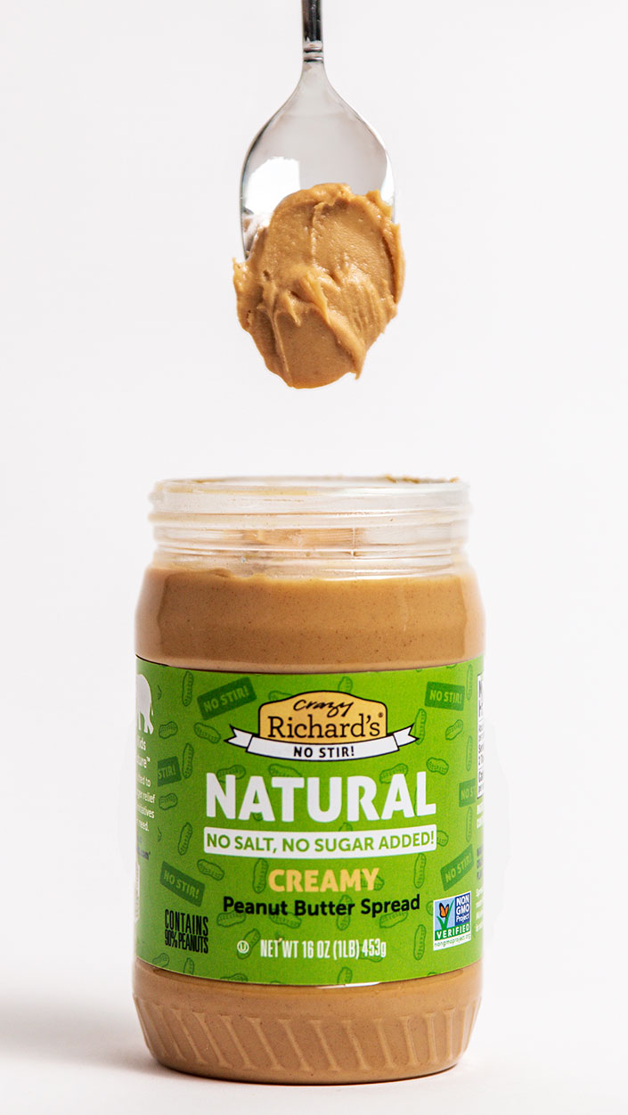 Photo of an open jar of Crazy Richard's Creamy Natural No-Stir peanut butter, with a spoon and a dollop of peanut butter suspended over the jar