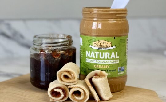 photo of a jar of Crazy Richards Natural No Stir Peanut butter, a jar of homemade jelly, and PB&J Sushi bites arranged on a wooden cutting board.