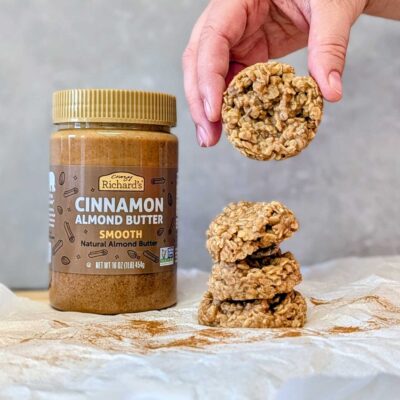 a hand holding a no-bake cookie over a stack of cookies on a counter. Behind the cookies is a jar of Crazy Richard's Cinnamon Almond Butter