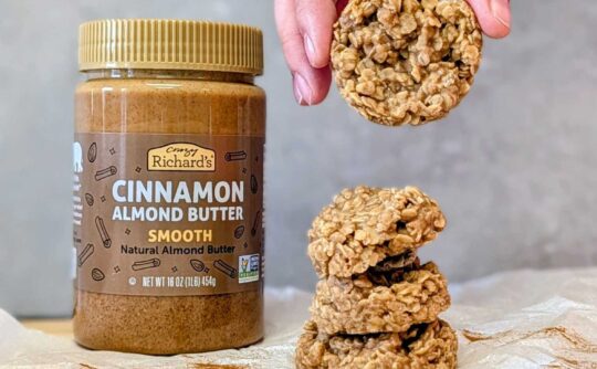 a hand holding a no-bake cookie over a stack of cookies on a counter. Behind the cookies is a jar of Crazy Richard's Cinnamon Almond Butter