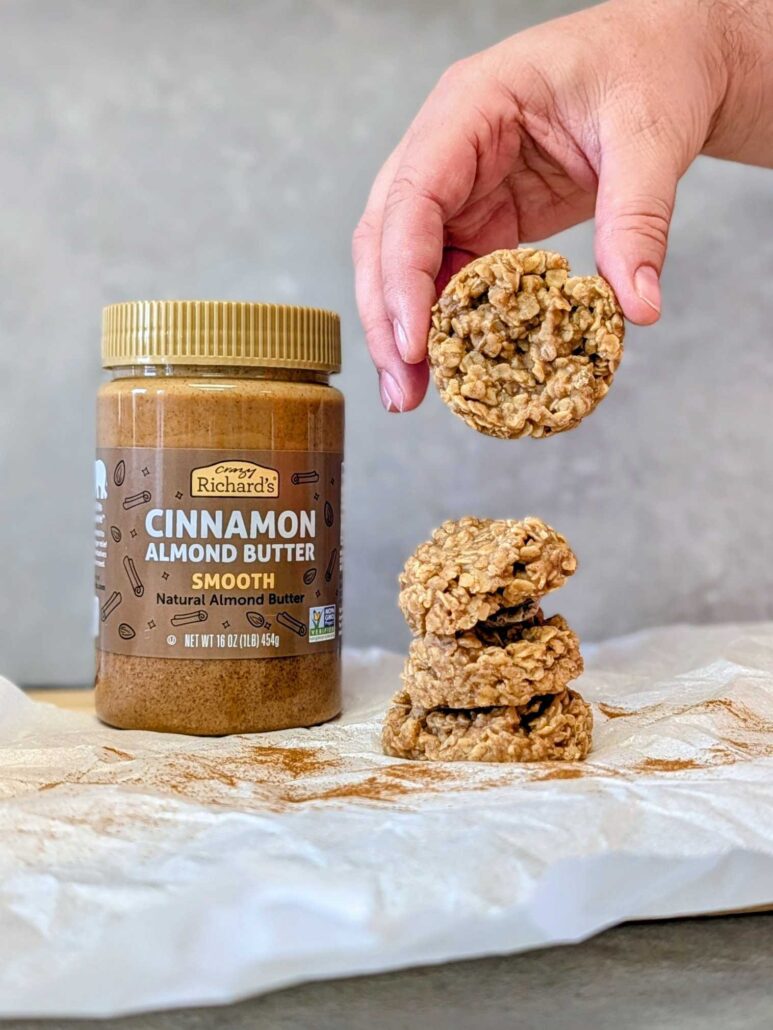 a hand holding a no-bake cookie over a stack of cookies on a counter. Behind the cookies is a jar of Crazy Richard's Cinnamon Almond Butter